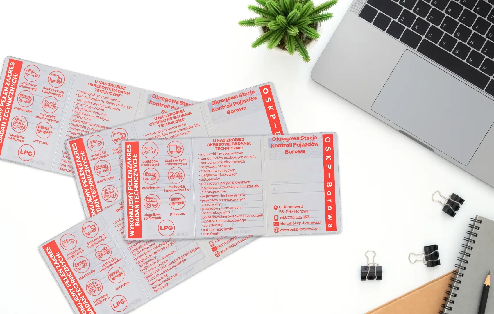 Cztery rozłożone okładki na dowody rejestracyjne z czerwonym nadrukiem stacji OSKP Borowa, leżące na białym biurku obok laptopa i roślinki.