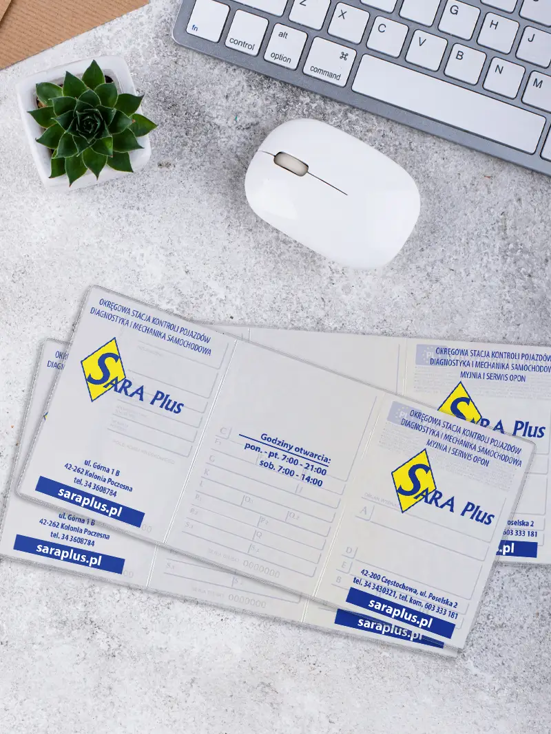 Zbliżenie na środkowe skrzydełko okładki na dowód rejestracyjny z nadrukiem godzin otwarcia stacji diagnostycznej, leżące obok białej myszki komputerowej.