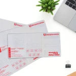 Cztery rozłożone okładki na dowody rejestracyjne z czerwonym nadrukiem agencji ubezpieczeniowej leżące na białym biurku obok laptopa.