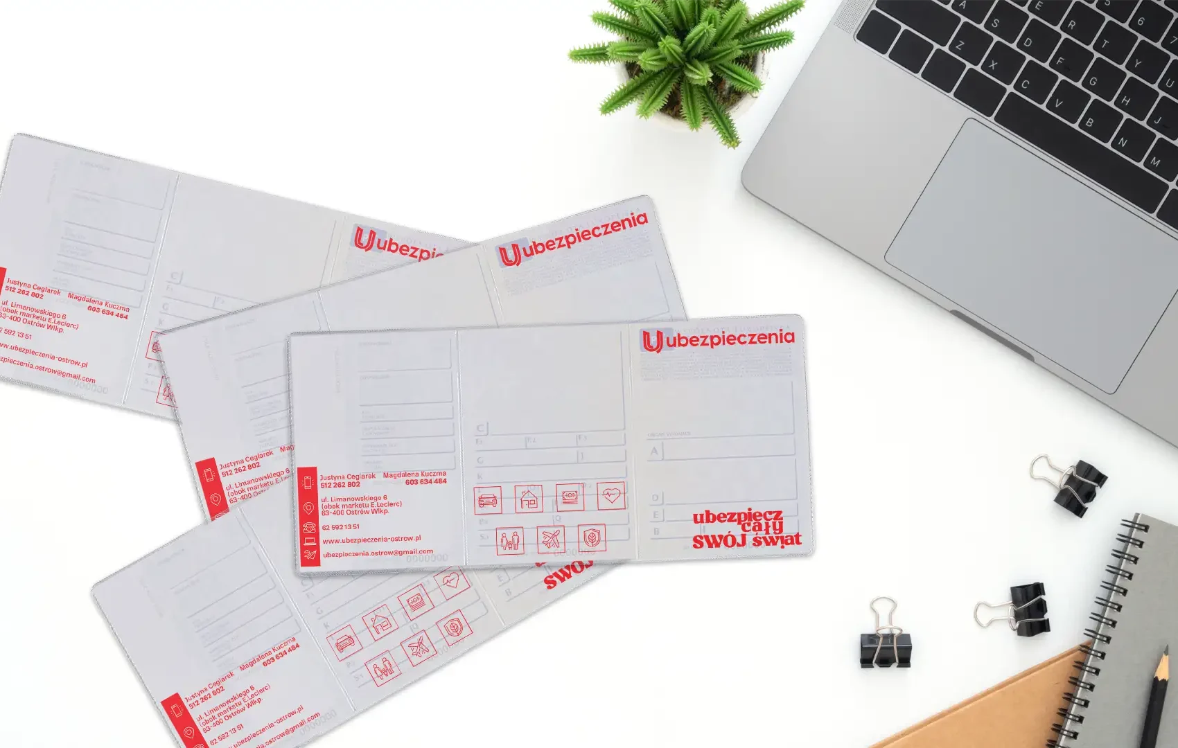 Cztery rozłożone okładki na dowody rejestracyjne z czerwonym nadrukiem agencji ubezpieczeniowej leżące na białym biurku obok laptopa.