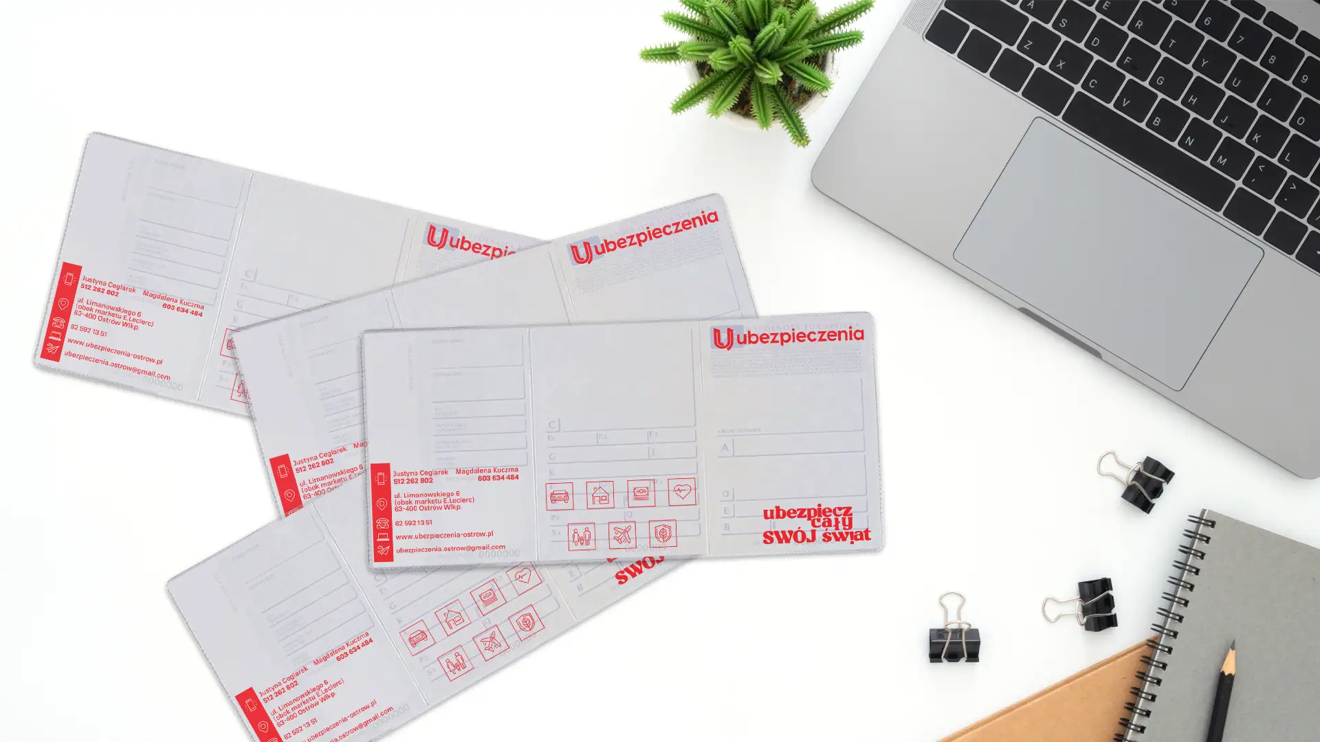 Cztery rozłożone okładki na dowody rejestracyjne z czerwonym nadrukiem agencji ubezpieczeniowej leżące na białym biurku obok laptopa.