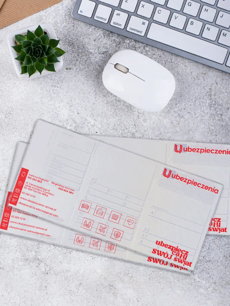 Zbliżenie na wnętrze okładki na dowód rejestracyjny z danymi kontaktowymi agenta i ikonami polis, ułożonej na szarym blacie obok białej myszki komputerowej.