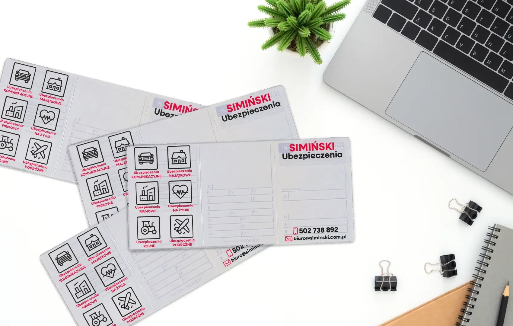Cztery rozłożone okładki na dowody rejestracyjne z czarnym nadrukiem agencji Ubezpieczenia Simiński leżące na białym biurku obok laptopa i notatnika.