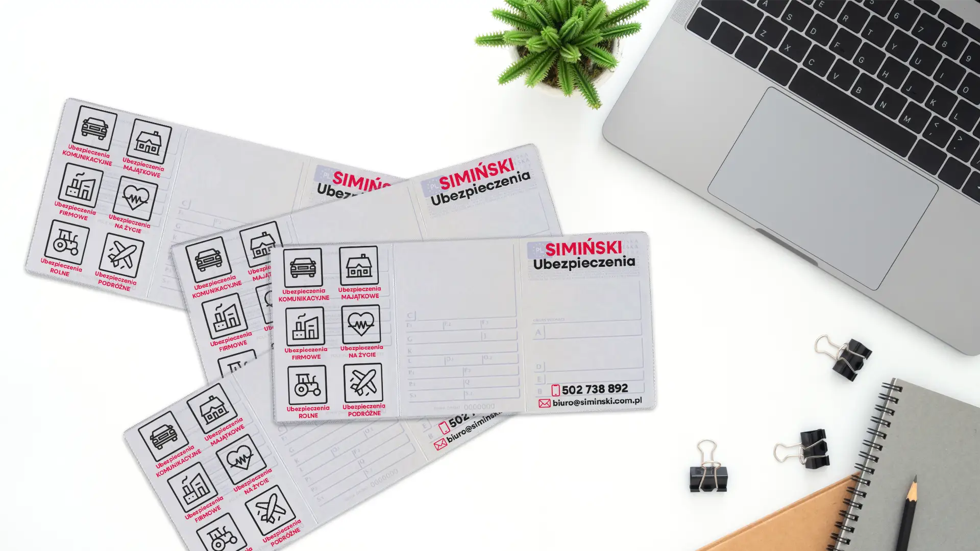 Cztery rozłożone okładki na dowody rejestracyjne z czarnym nadrukiem agencji Ubezpieczenia Simiński leżące na białym biurku obok laptopa i notatnika.